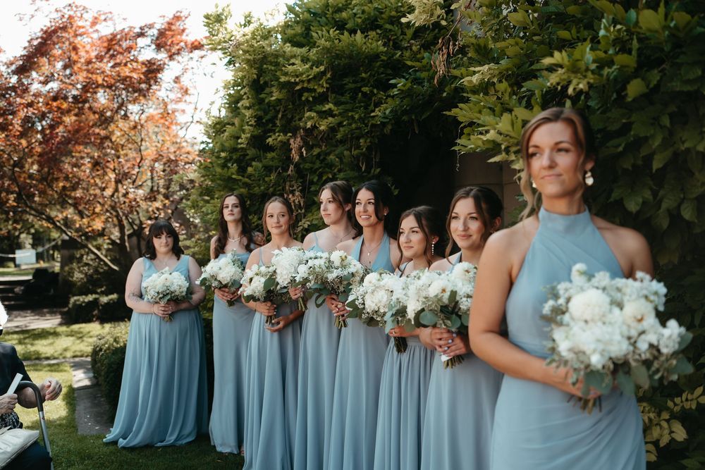 Row of bridesmaids in matching light blue gowns holding white floral bouquets at outdoor wedding ceremony.