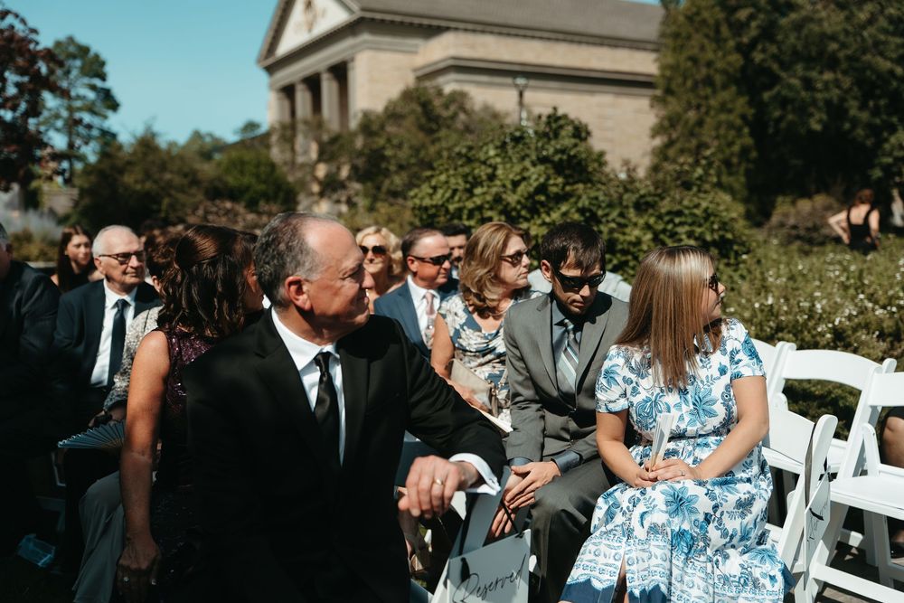 Wedding guests seated outdoors in white chairs enjoying a sunny ceremony near a historic stone building.