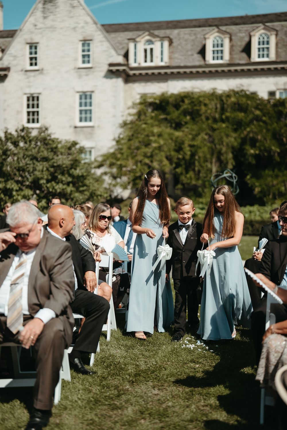 Flower girls in blue dresses and ring bearer walk down outdoor wedding aisle at elegant stone manor.