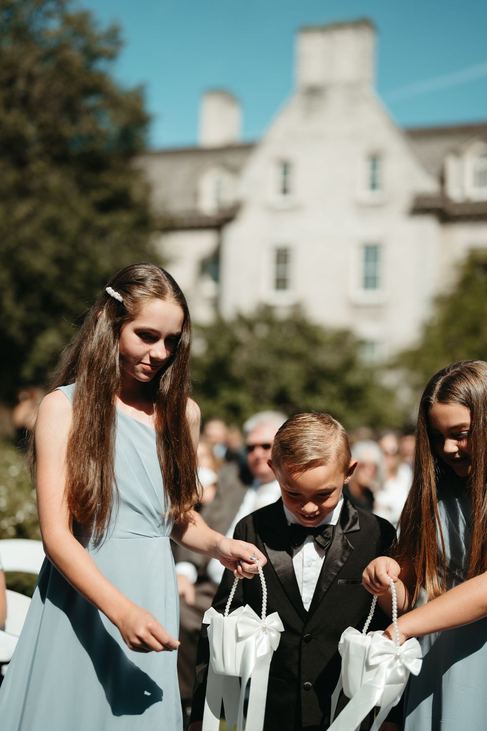Two flower girls in blue dresses and a ring bearer in a tuxedo hold baskets at an outdoor wedding ceremony.