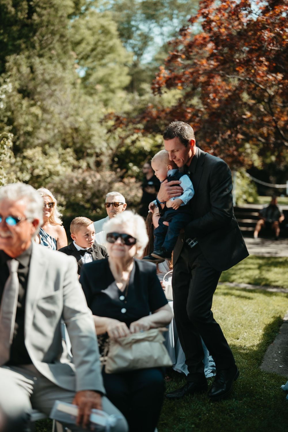 A man in a black suit holds a baby at an outdoor wedding ceremony surrounded by seated guests.