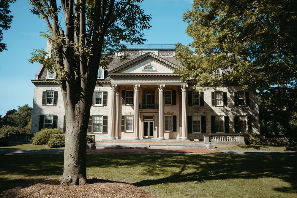 Elegant neoclassical mansion with white columns, symmetrical facade, and lush green lawn surrounded by mature trees on a sunny day.