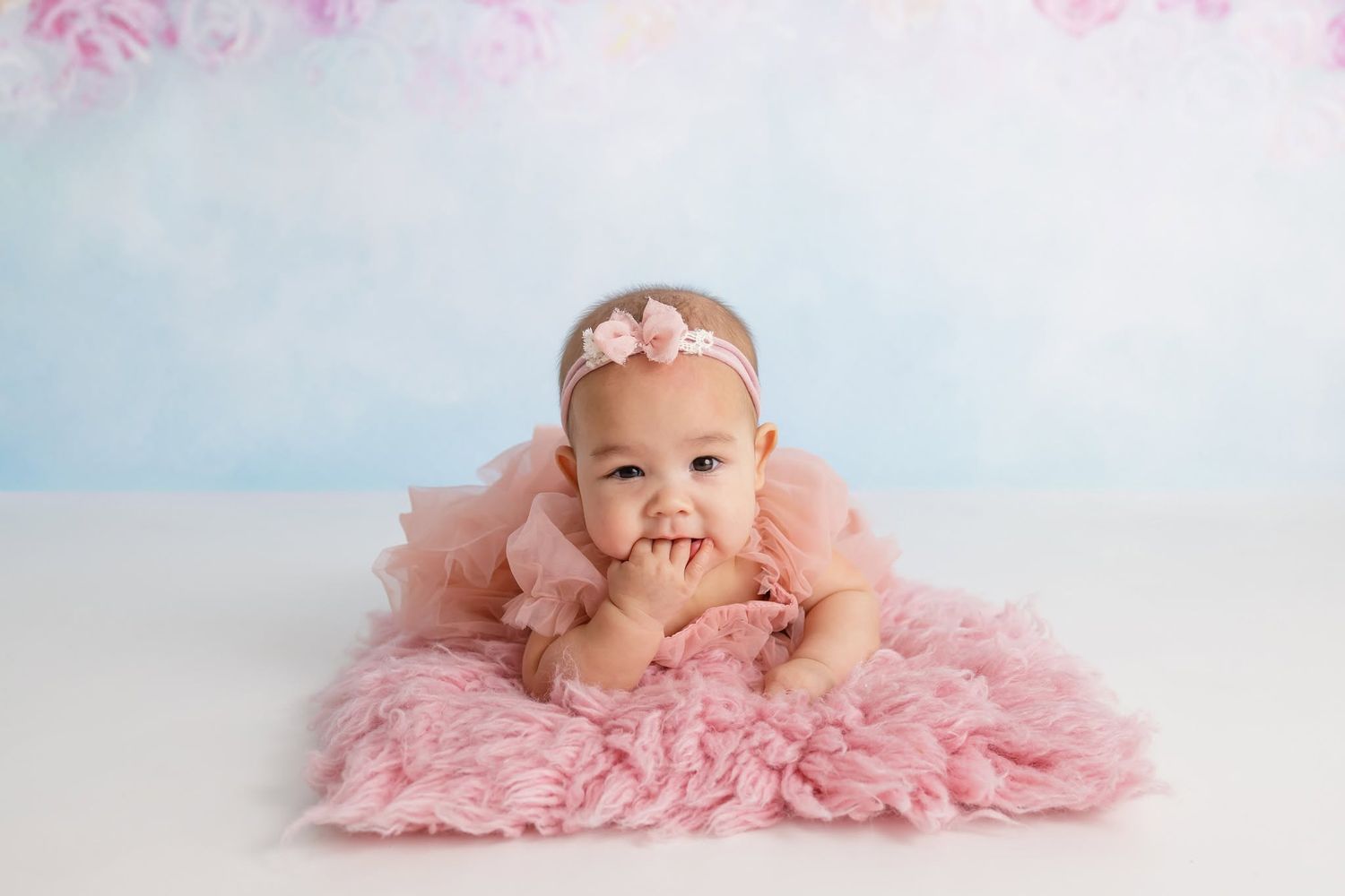 Baby girl in pink ruffle dress and floral headband sits on fluffy white rug in bright studio setting.