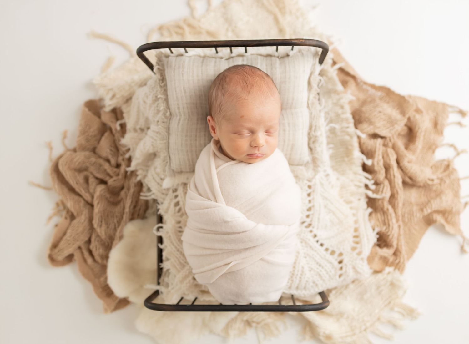 Newborn baby swaddled in white wrap sleeping peacefully in a small metal bed with cream and burlap textured blankets.