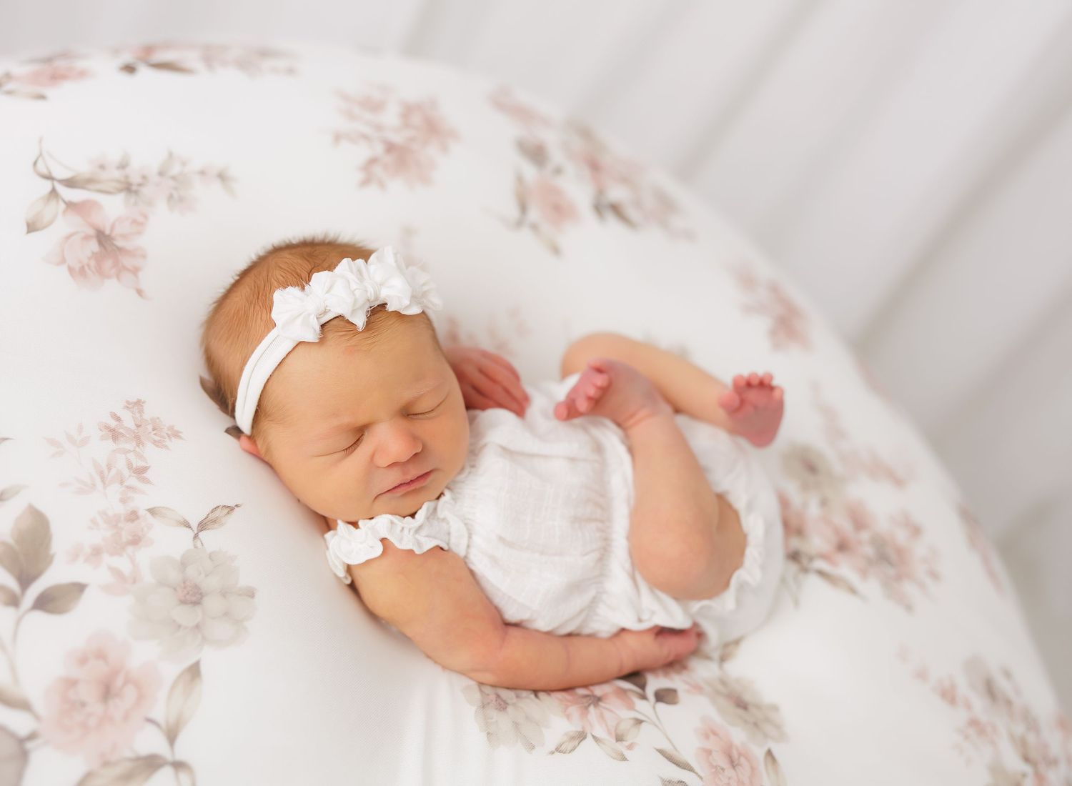 Sleeping newborn baby girl in white romper and floral headband on a soft floral beanbag.