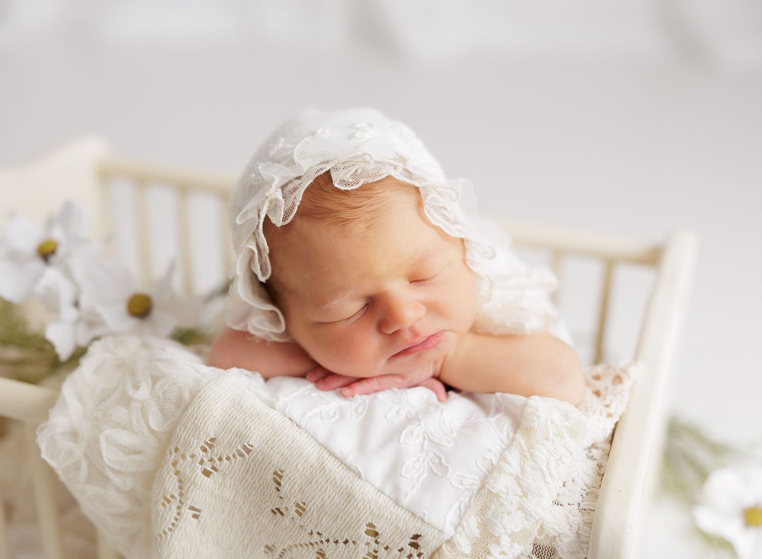 Peaceful newborn baby sleeping in a white lace bonnet and blanket in a tiny wooden bed with flowers.
