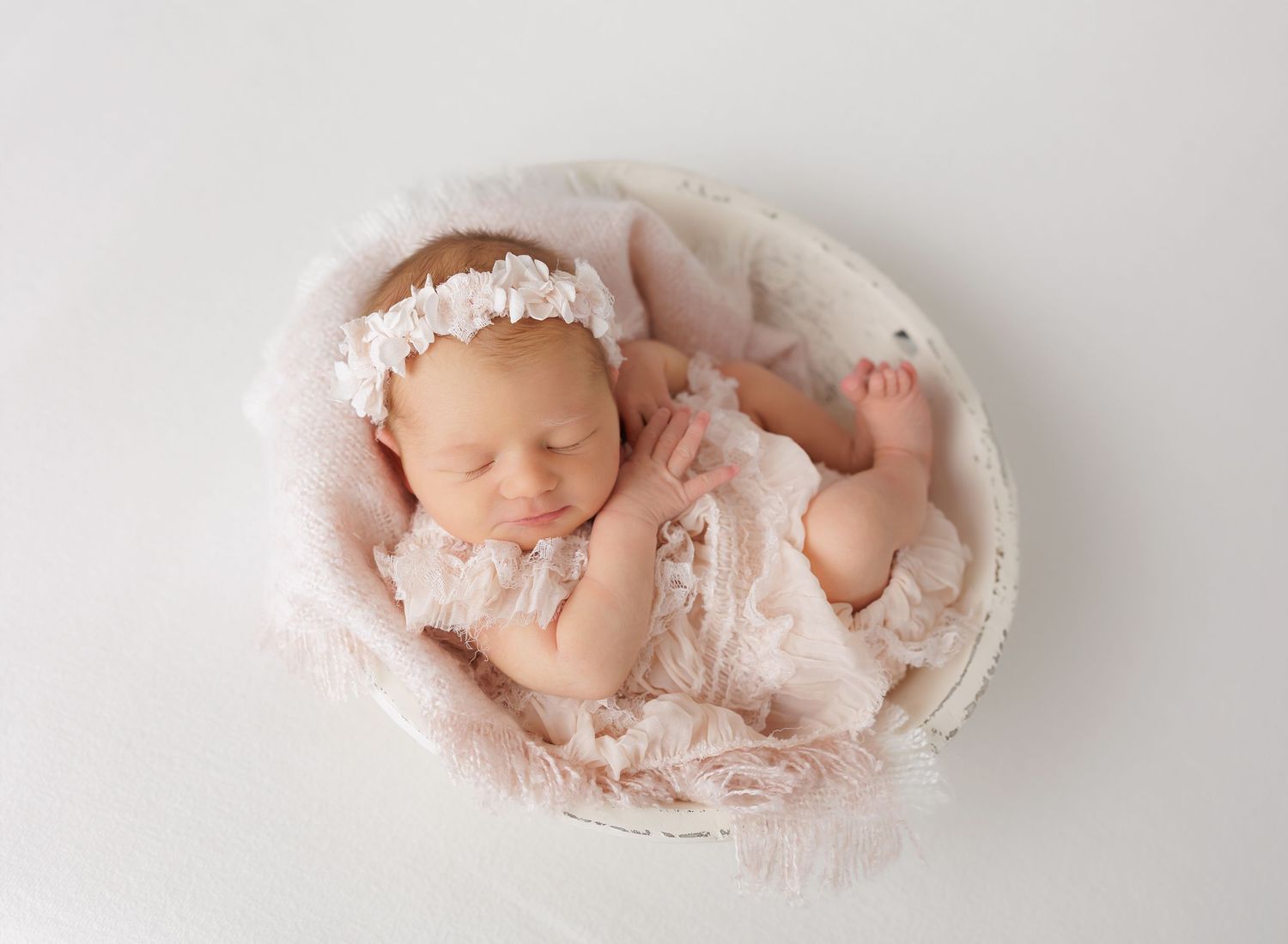 Sleeping newborn baby girl in pink lace outfit resting in a white bowl with soft pink fluff.