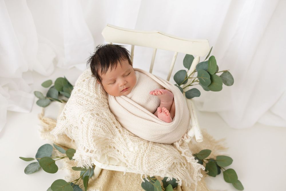 Newborn baby swaddled in cream wrap sleeping in a bowl prop surrounded by eucalyptus leaves on white background.