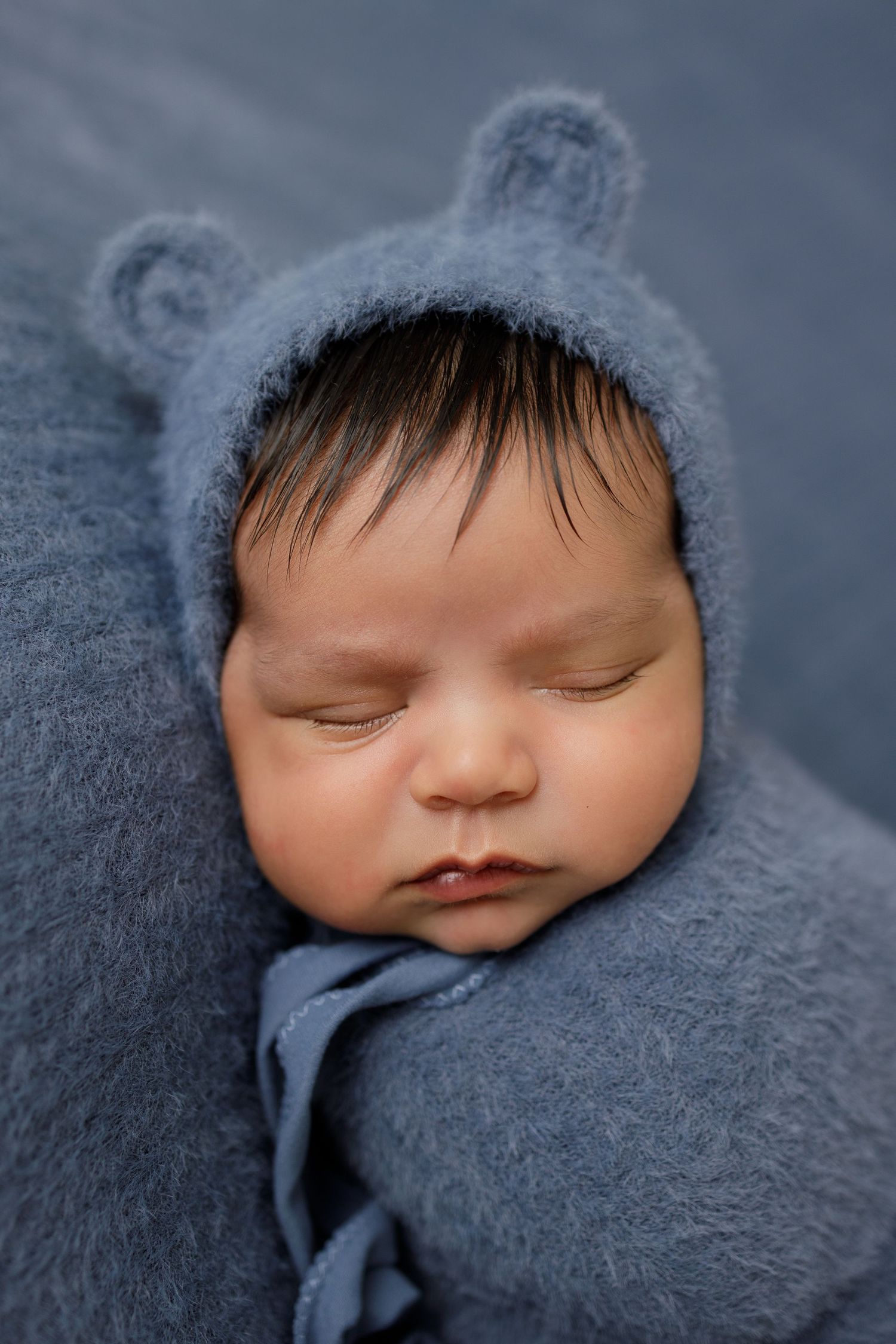 Sleeping newborn baby wrapped in blue knit blanket wearing a bear ear bonnet, peaceful infant portrait.