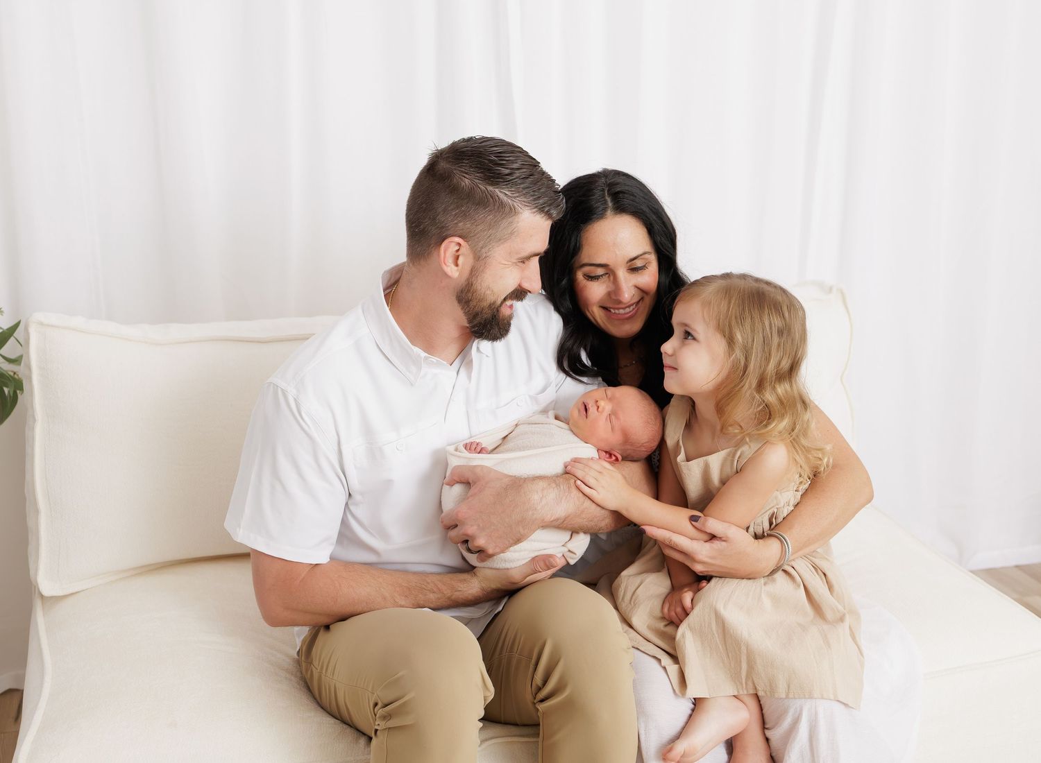 Happy family of four sitting on white sofa, parents smiling at newborn baby while young daughter reaches out to touch sibling.