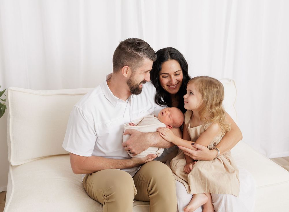 Happy family of four sitting on white sofa, parents smiling at newborn baby while young daughter reaches out to touch sibling.