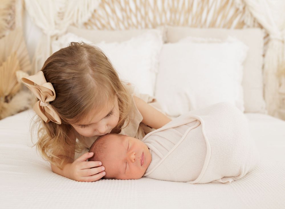Young girl with bow gently kisses sleeping swaddled newborn baby sibling on white bed.