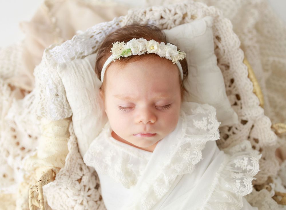Sleeping newborn baby wearing a delicate white floral headband, wrapped in lace blankets for a soft, elegant portrait.