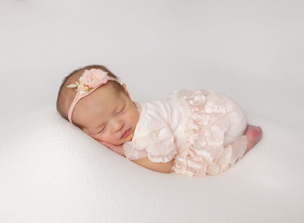 Sleeping newborn baby girl in pink lace romper and floral headband posed on white backdrop.