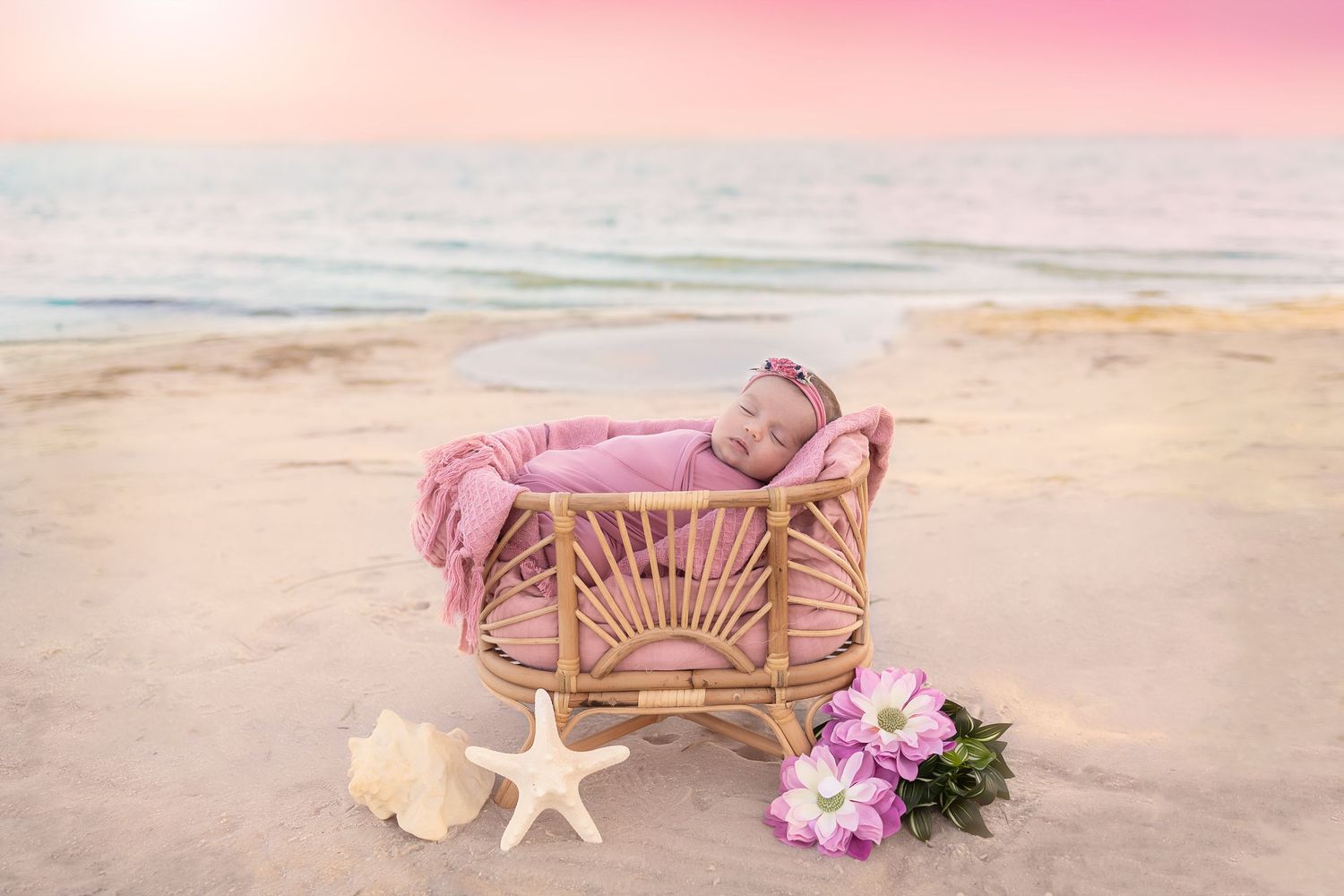Newborn baby sleeping in a wicker bassinet on a sandy beach at sunset with pink blanket and flowers.