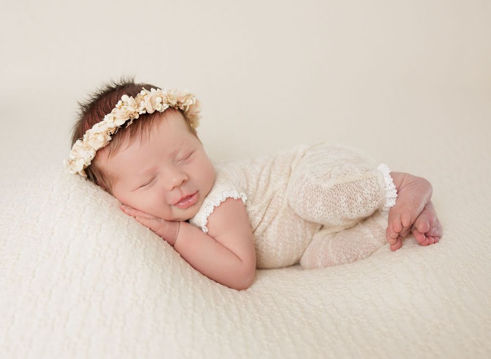 A sleeping newborn baby wearing a floral headband poses peacefully in a cream lace romper on a white blanket.