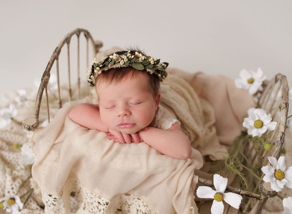 Sleeping newborn baby wearing a floral crown resting in a rustic mini bed with white cosmos flowers.