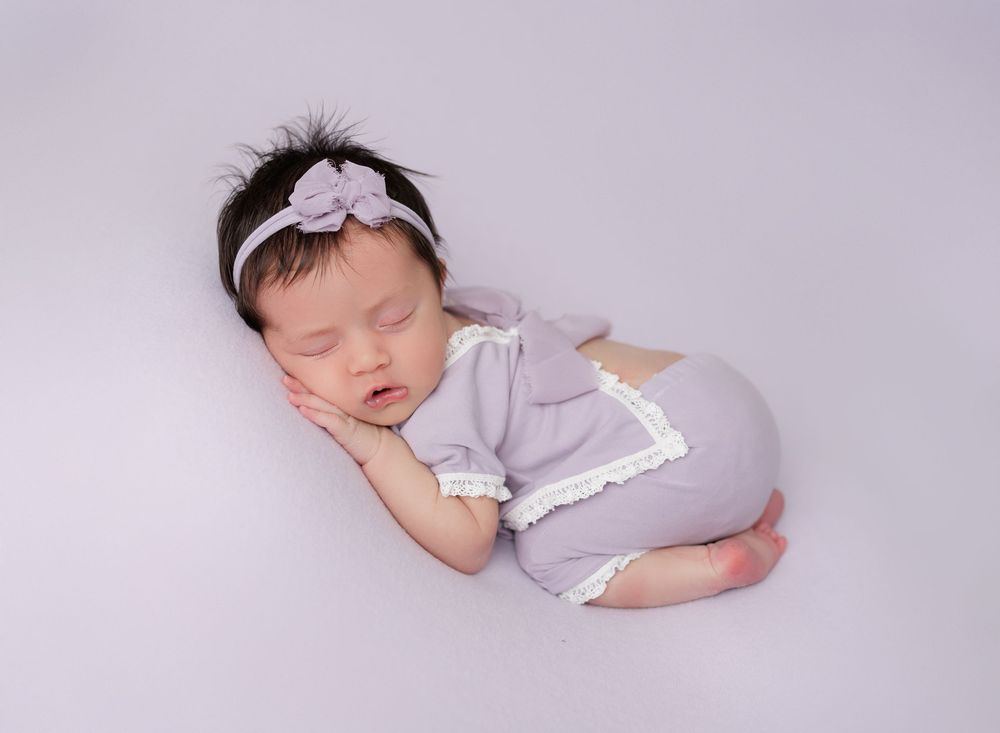 Sleeping newborn baby in lavender romper with lace trim and floral headband on soft gray background.
