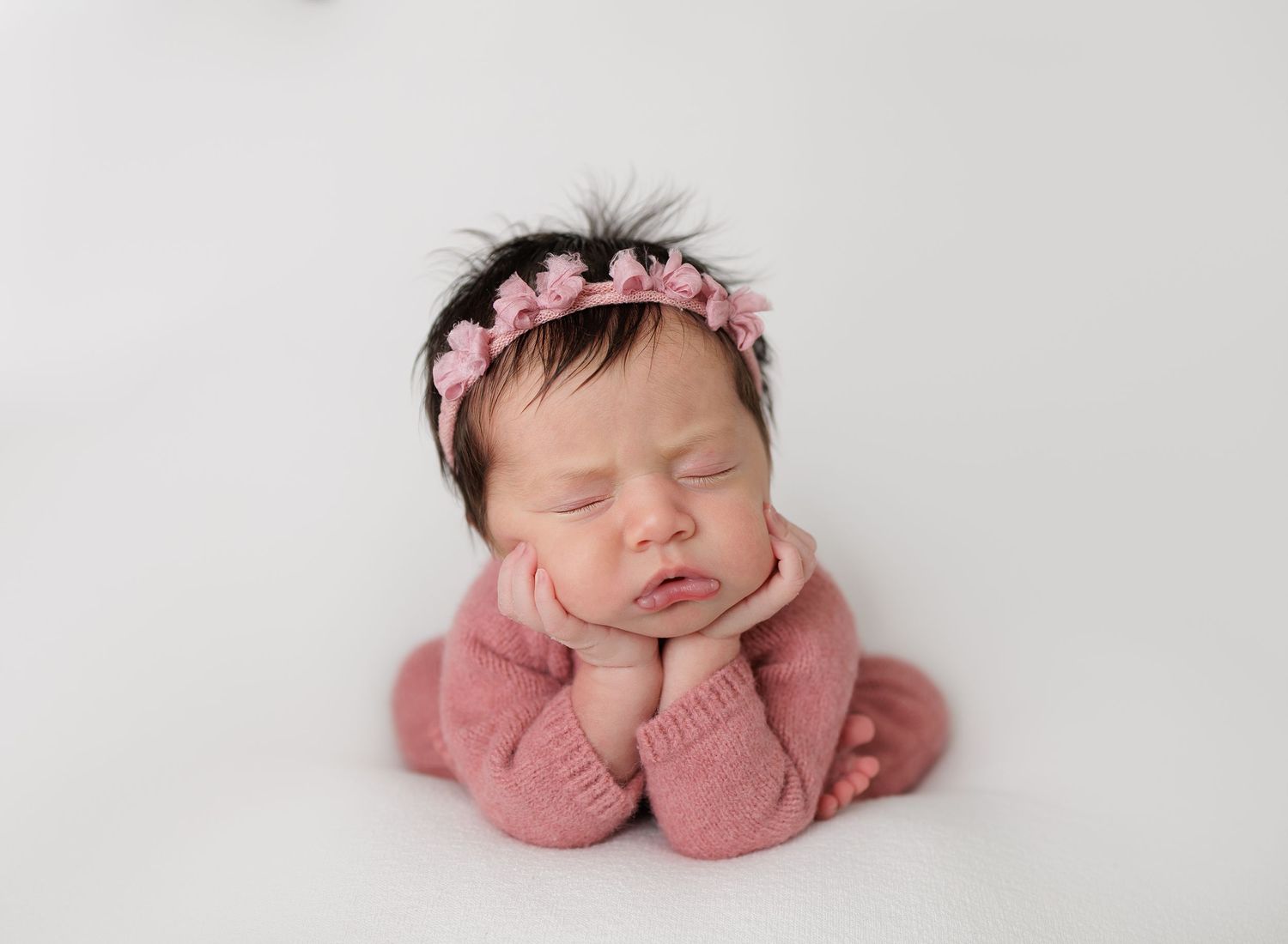 Sleeping newborn baby girl in pink knit outfit and floral headband posed with hands under chin on white background.