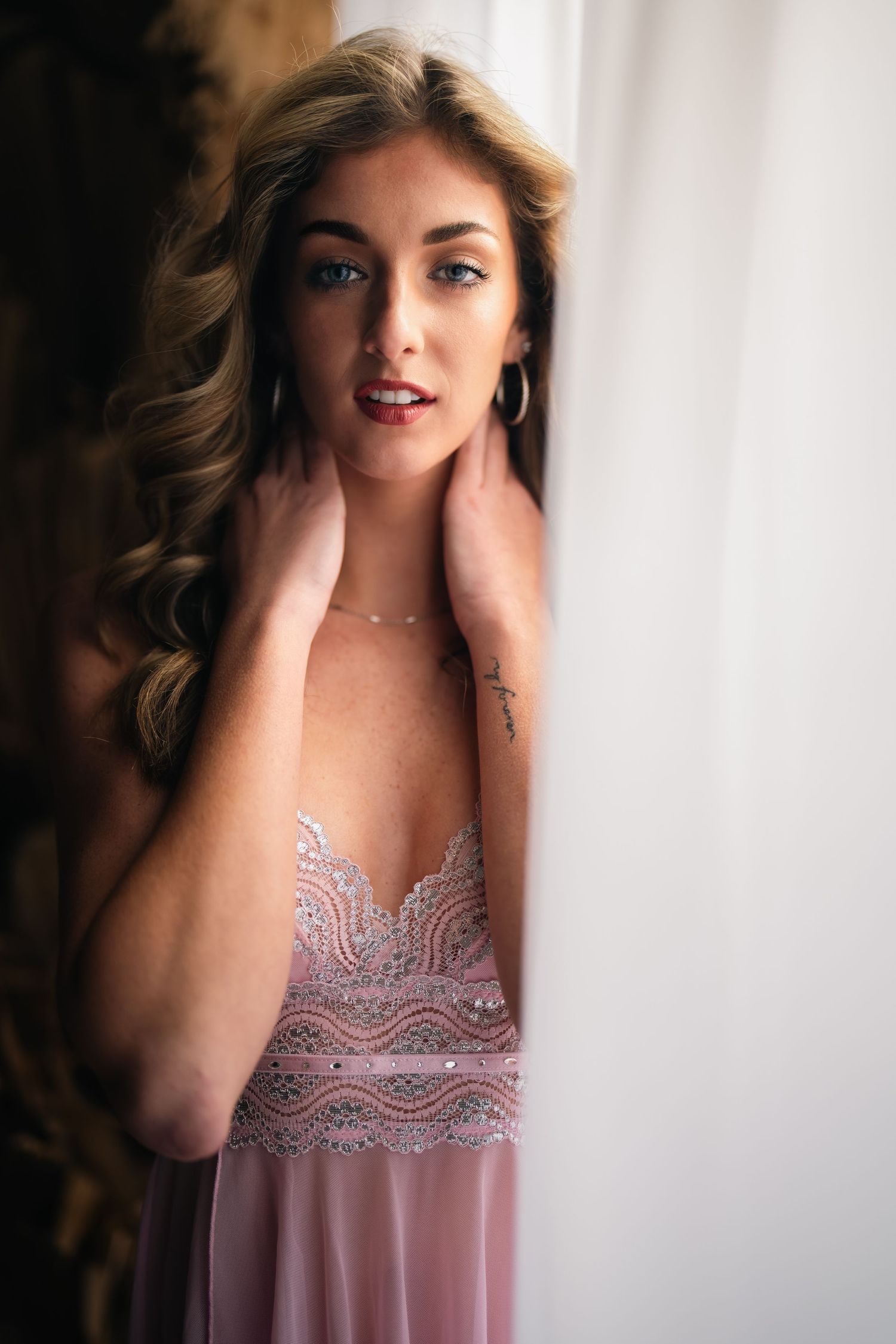 Woman in pink beaded gown posing elegantly near a window with soft natural light highlighting her features.