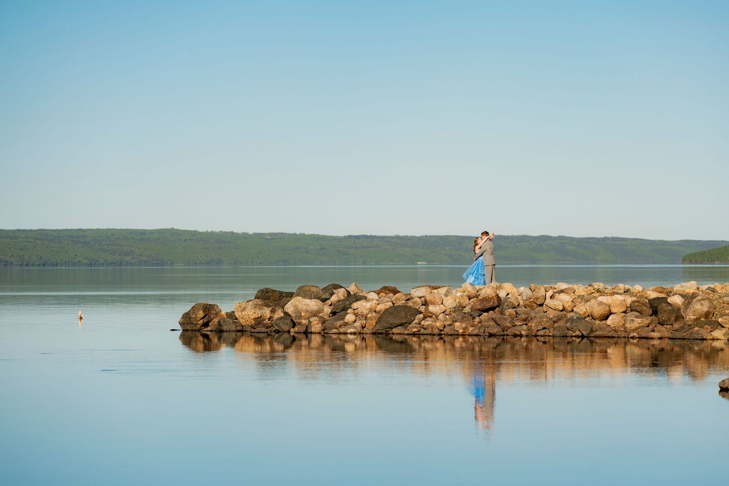 Person standing on rocky jetty fishing in calm reflective lake with green hills and clear blue sky.
