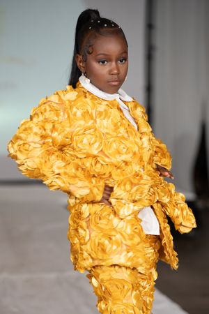 Young girl model wearing a golden yellow 3D rosette textured suit on a fashion runway.