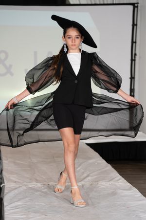 Young model walks runway in black blazer, shorts, sheer cape, and wide-brim hat at fashion show.