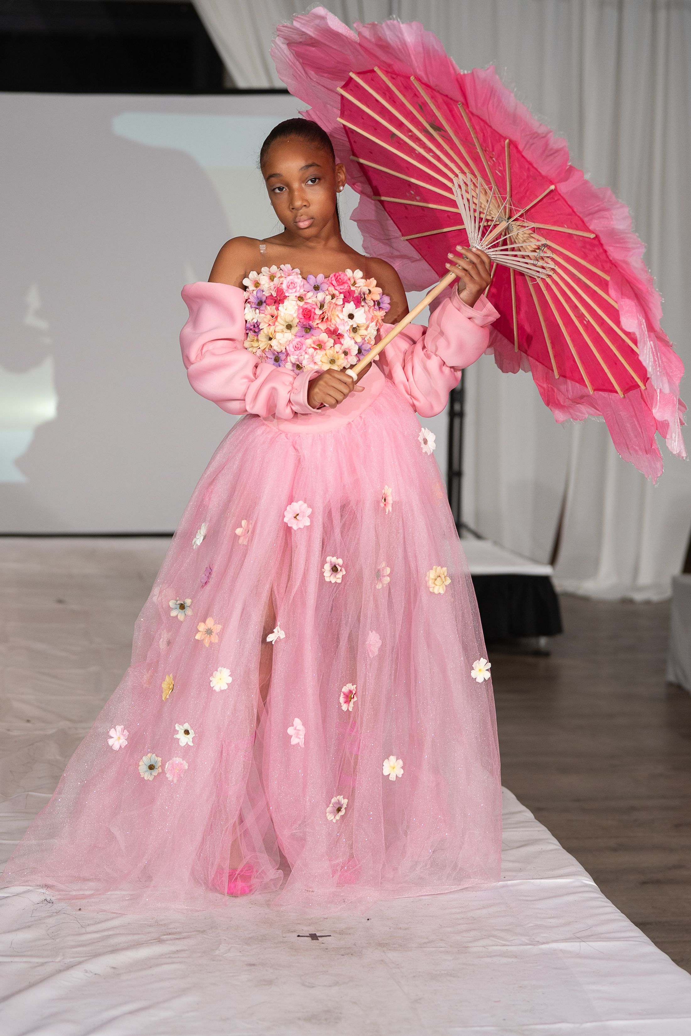 Young girl in pink floral tulle gown holding a pink parasol on a fashion runway.