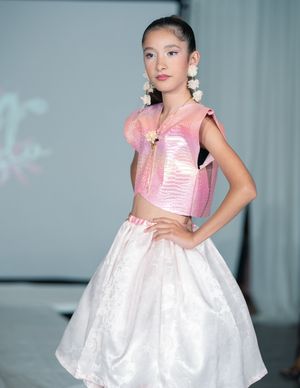 Young girl model on runway wearing pink iridescent crop top and white floral skirt with floral earrings.