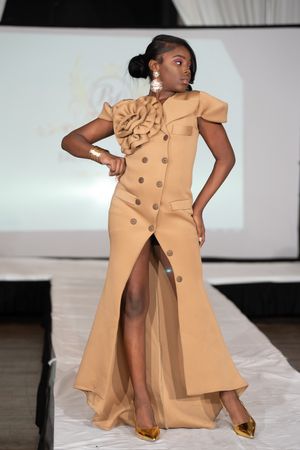 Model walks runway in elegant tan button-front gown with ruffle detail and gold heels at fashion show.