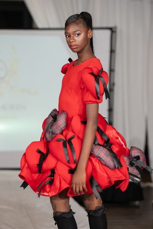 Young model on runway wearing red dress with black bows and plaid accents at fashion show.