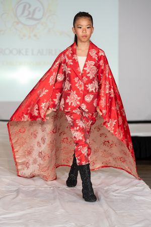 Young model walks runway in red floral brocade suit with matching cape at Brooke Laure fashion show.