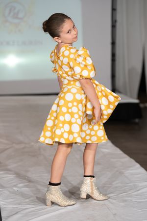 Young girl modeling a yellow polka dot dress with white ankle boots on a fashion runway.