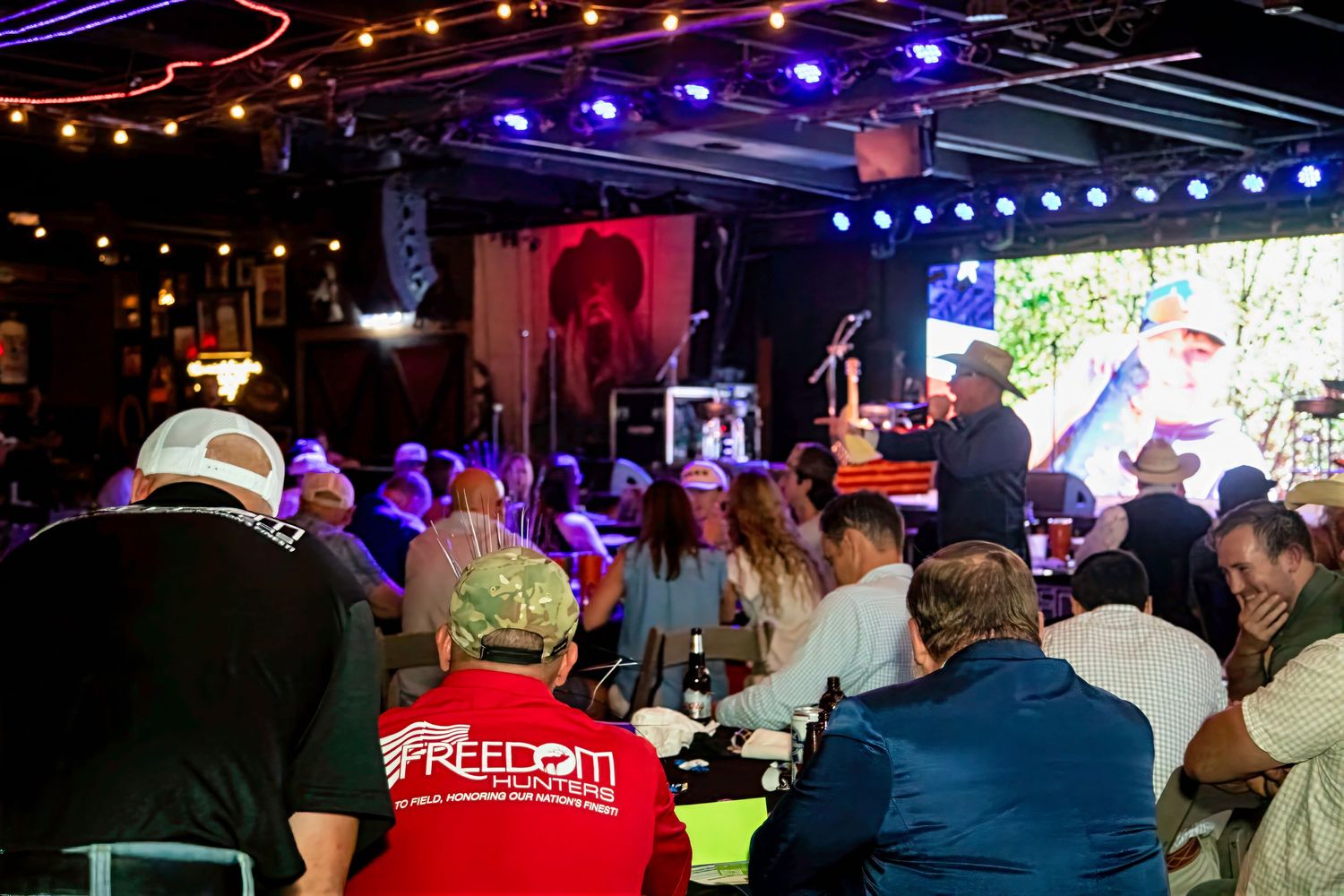 Crowd enjoying a live country music performance at an outdoor bar venue with stage lighting and large video screens.