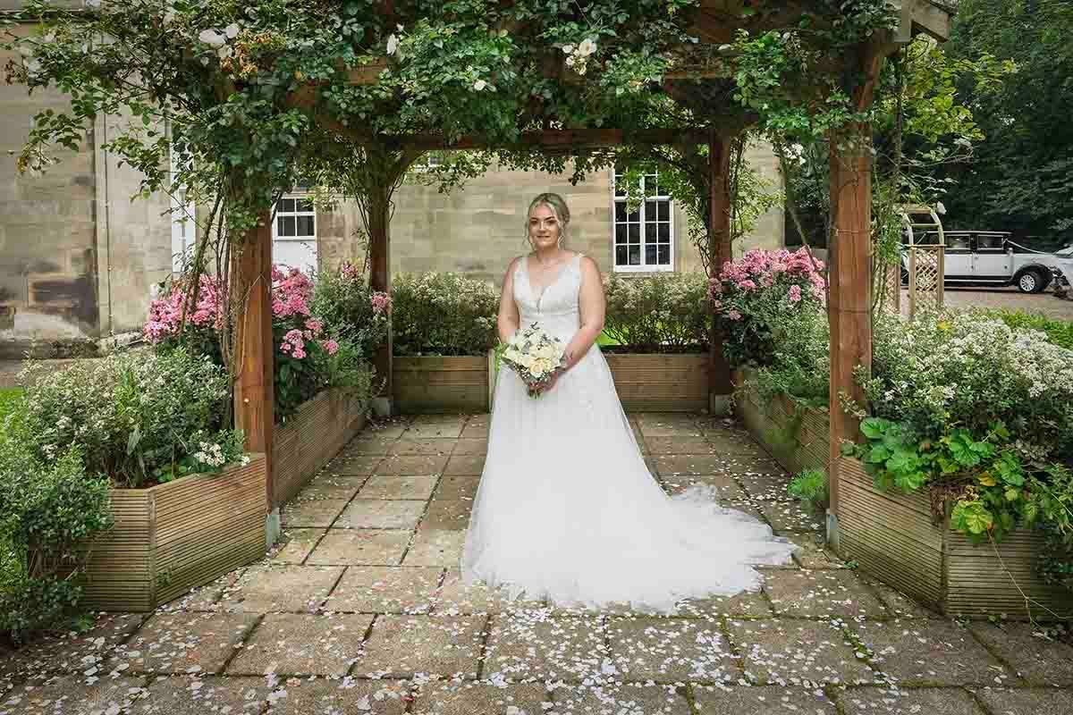 Bride in white tulle gown holding bouquet under wooden pergola surrounded by lush garden flowers at Lady Gillfords House in Carlisle