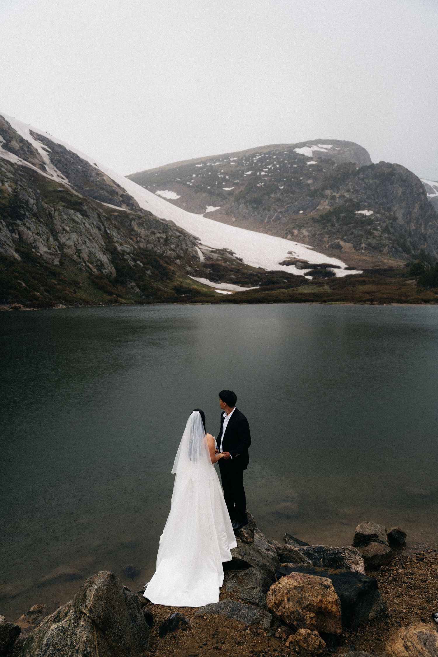 Bride and groom standing on rocky shore of alpine lake with snow-capped mountains in misty background.