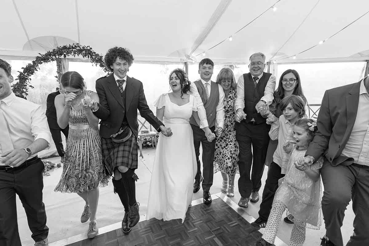 Black and white wedding photo of bride and groom dancing joyfully with guests in a marquee venue.