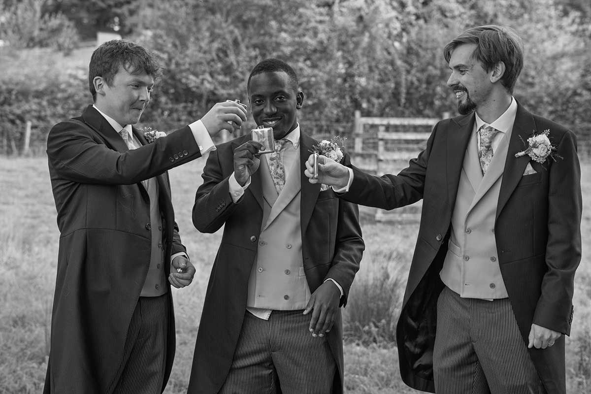 Three men in formal suits toasting with drinks at a wedding celebration in a black and white photo.