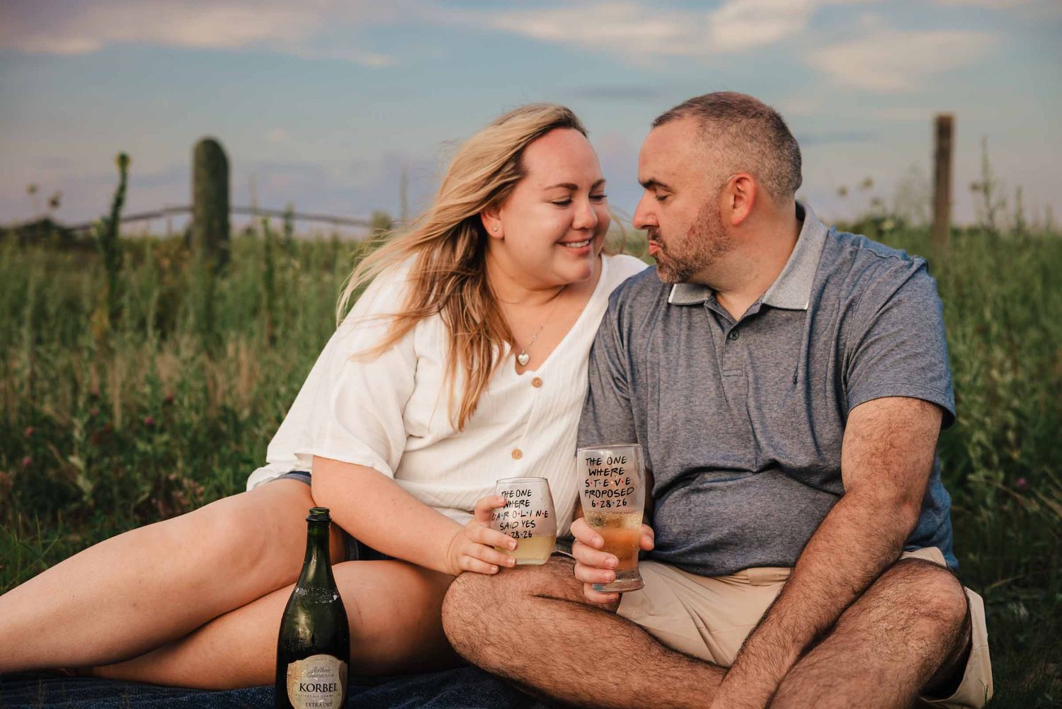 Engaged couple sitting together holding personalized champagne glasses during a picnic near Duxbury Beach.