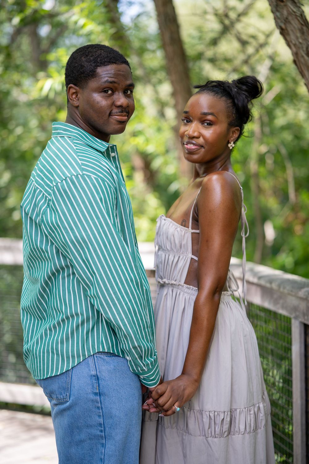 A young Black couple holds hands on a wooden bridge surrounded by lush green trees, smiling at the camera.