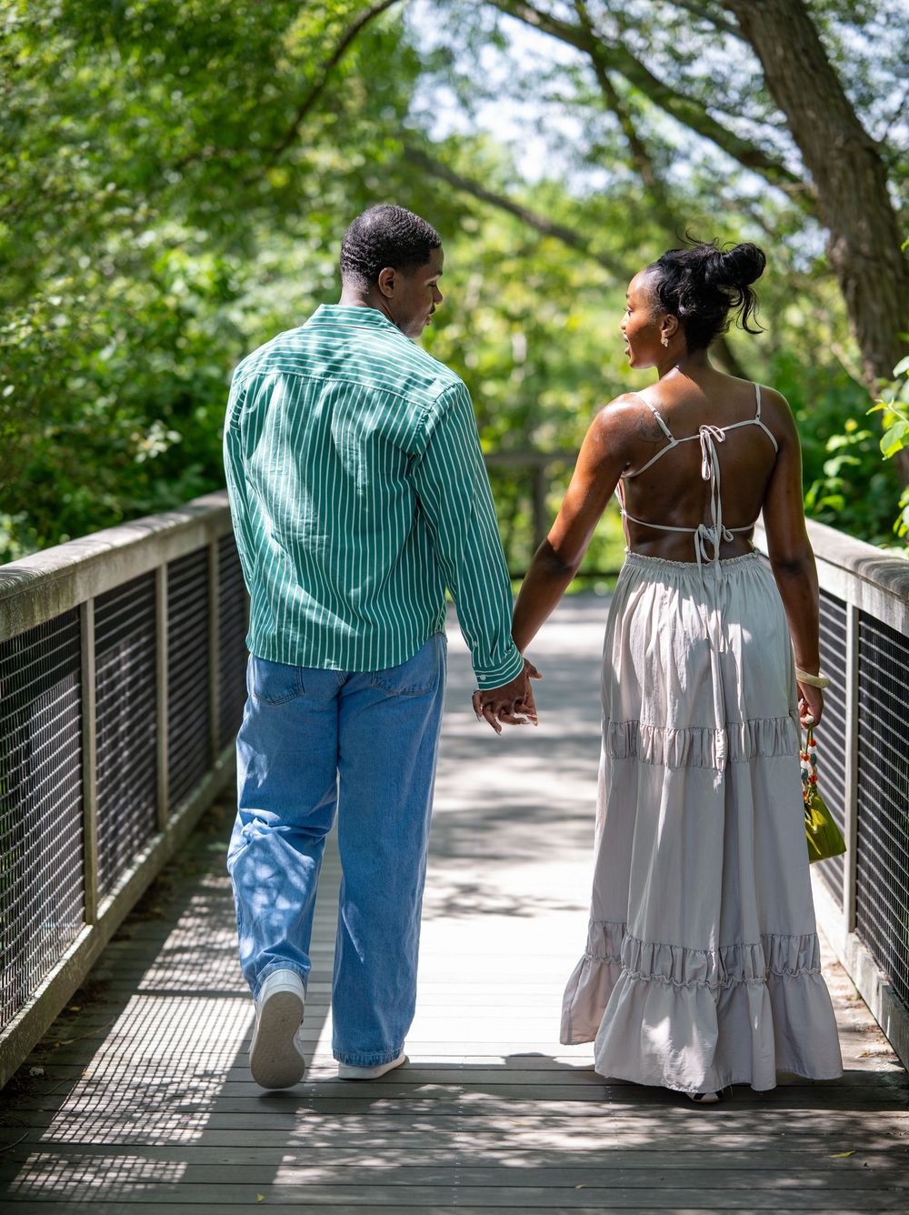 A Black couple walks hand-in-hand on a wooden bridge surrounded by lush green trees on a sunny day.