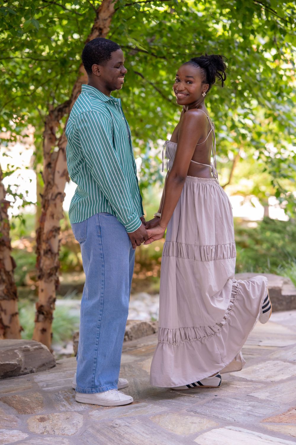 A young Black couple holds hands and smiles while walking through a lush green park, dressed in casual summer attire.