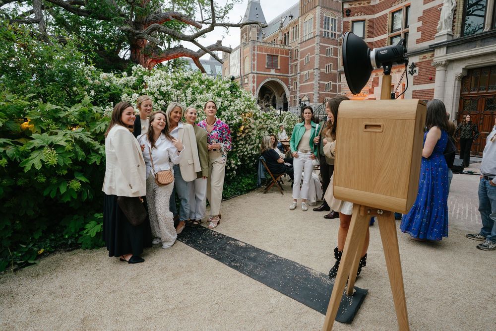 Group of people posing for a large format camera on a tripod outdoors near a historic brick building with blooming trees.