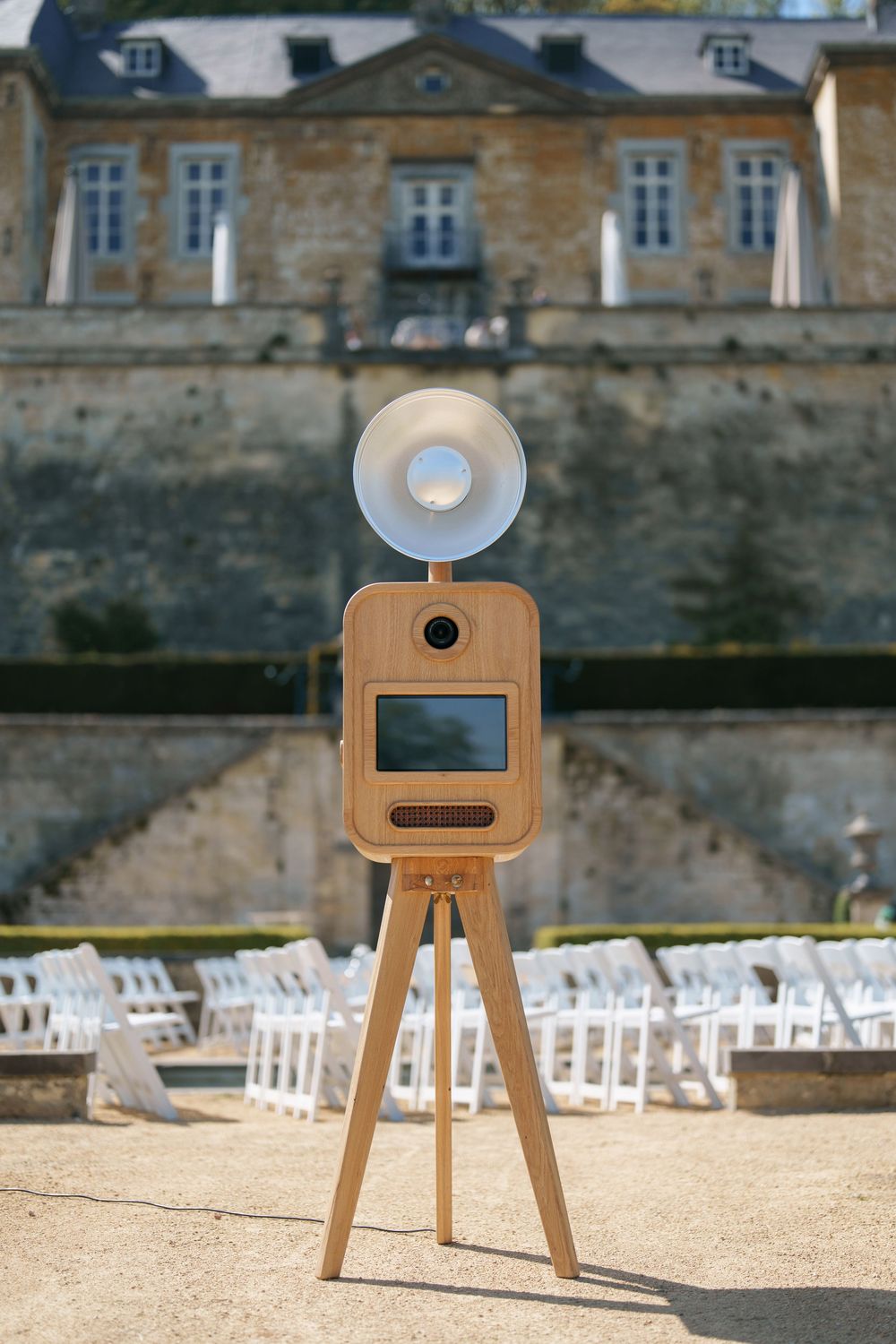 Modern wooden photo booth on tripod with ring flash at outdoor wedding venue with white chairs and historic building.