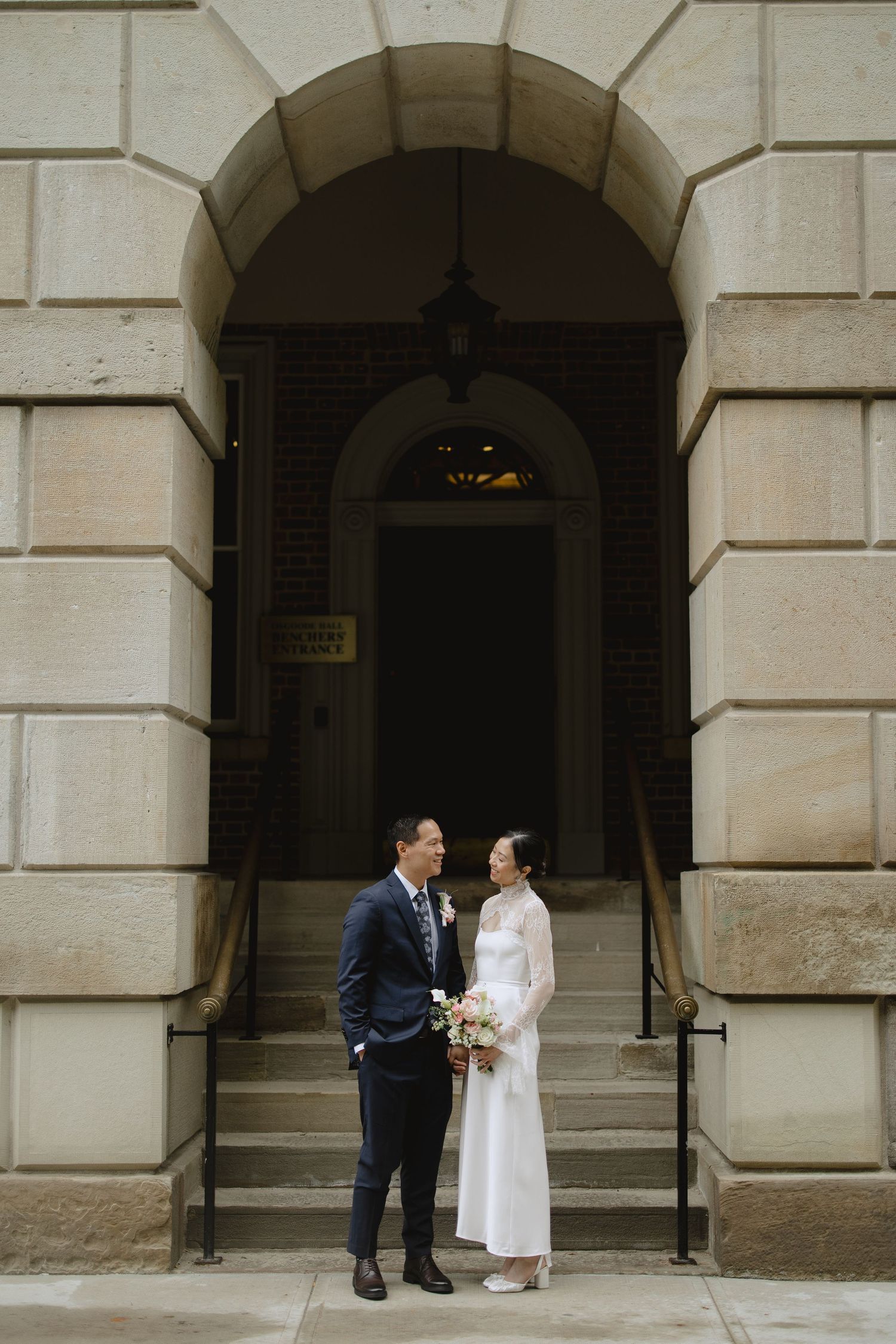 A bride in a white dress and a groom in a navy suit stand together on stone steps beneath a grand arched entrance.
