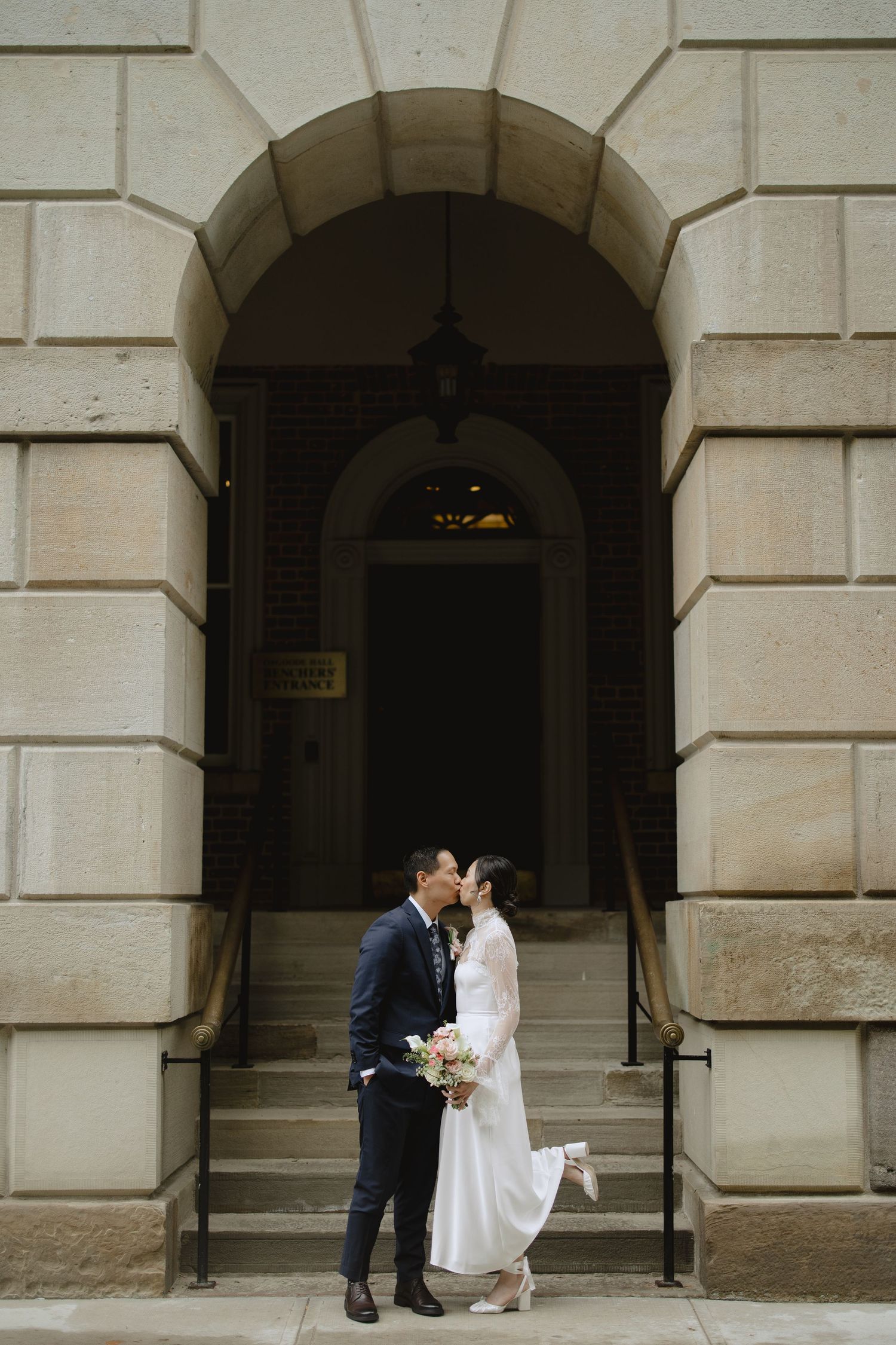 A bride in a white gown holding a bouquet kisses her groom in a navy suit beneath a grand stone archway with steps behind them.