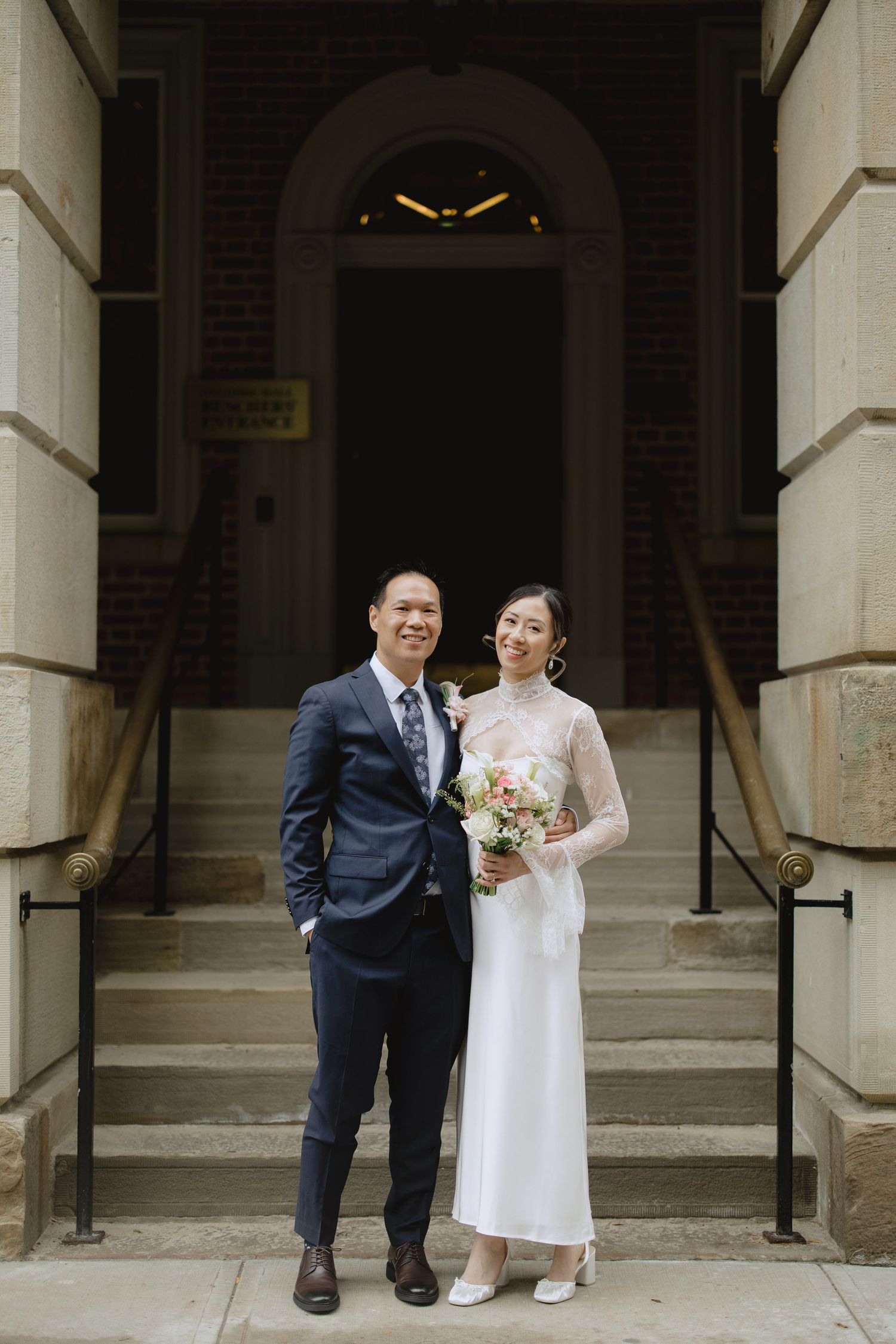 Smiling bride in a white long-sleeve dress holding a pink bouquet stands beside a groom in a navy suit on brick building steps.