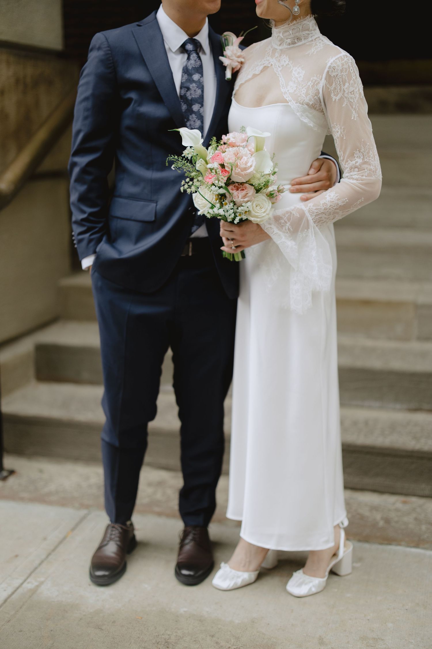 Bride in a white lace-sleeved satin wedding dress holding a pink floral bouquet stands beside a groom in a navy suit on stone steps.