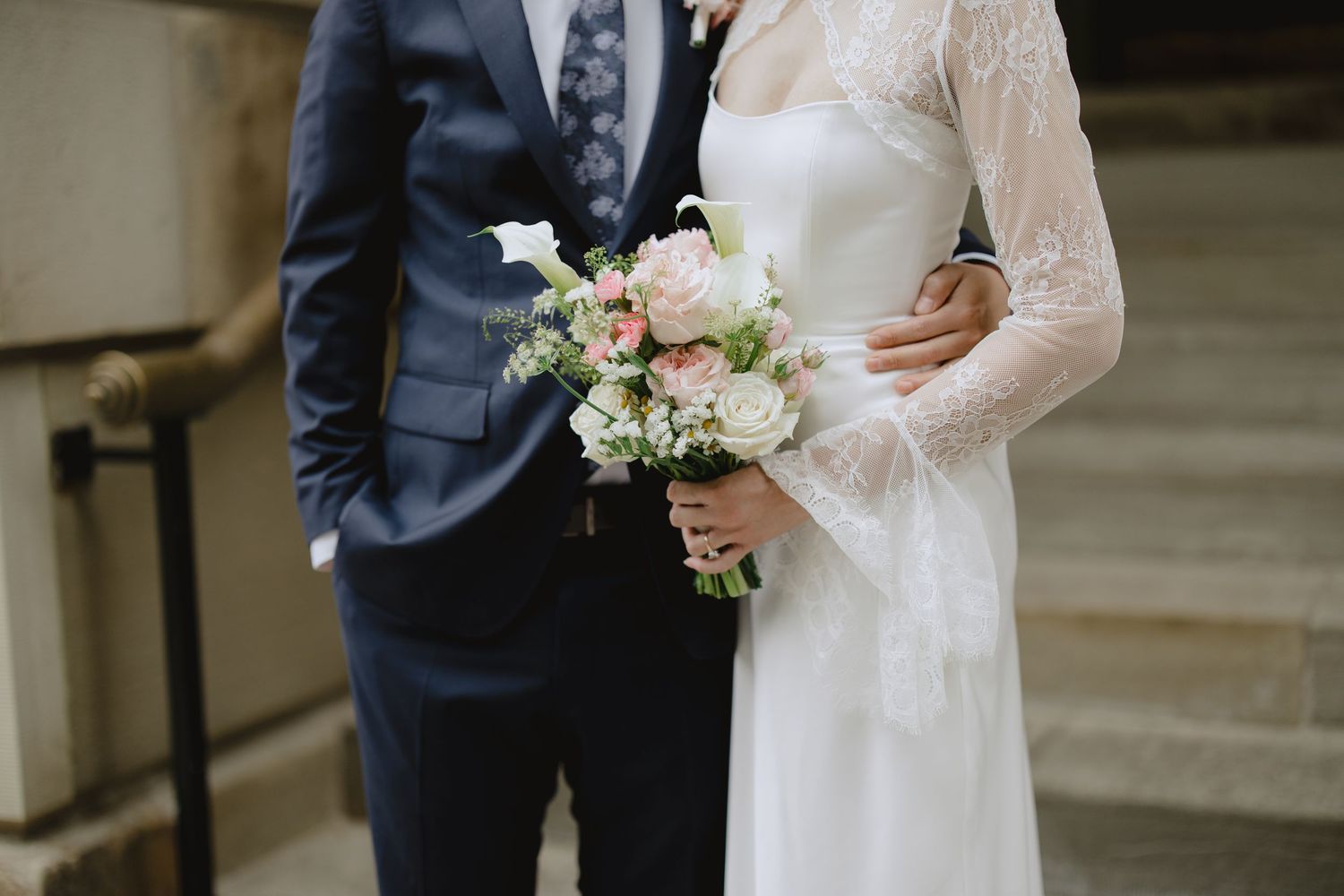 Bride in a white lace wedding dress holding a pink and white bouquet stands beside a groom in a navy suit on stone steps.