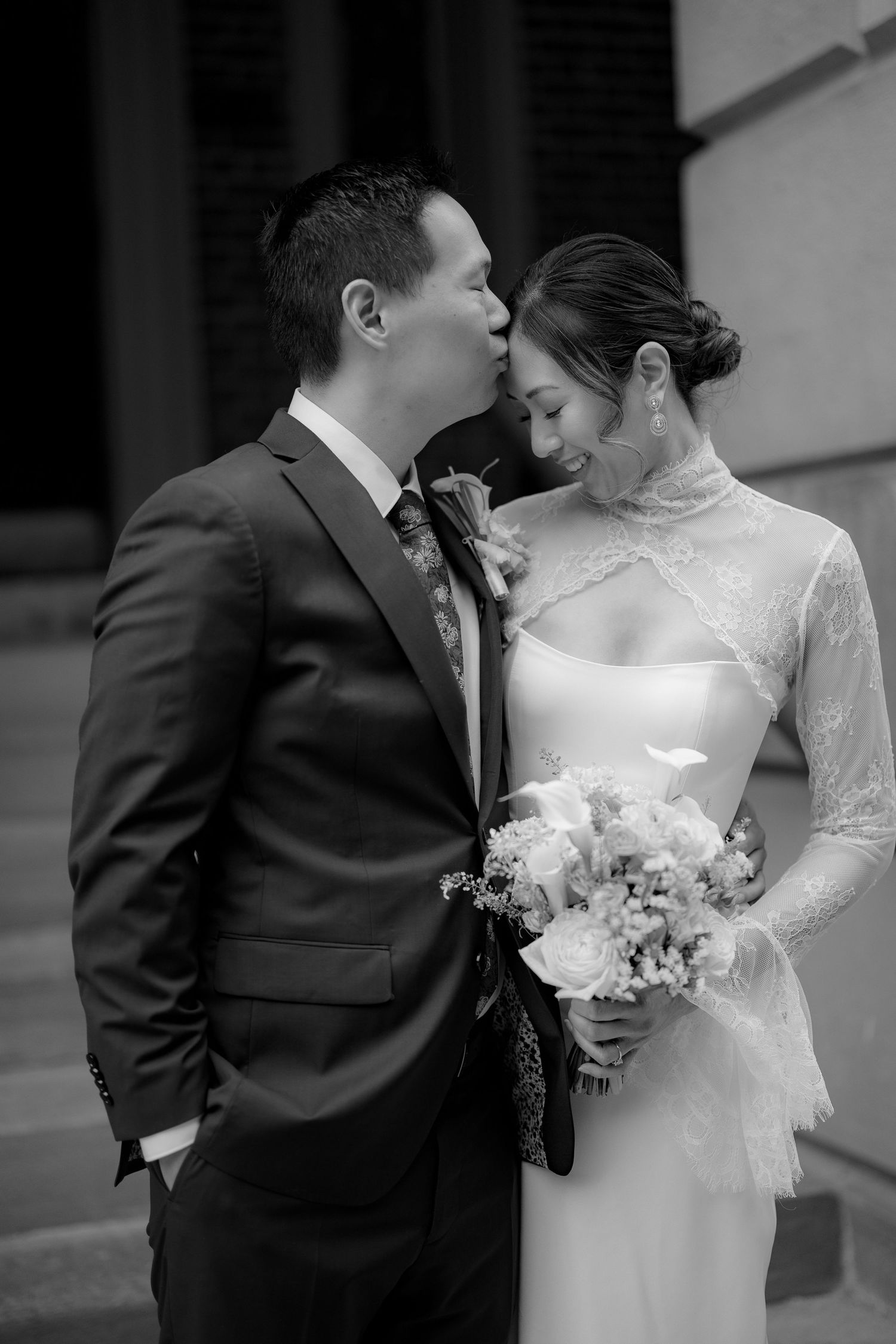 Black and white wedding portrait of a groom in a suit kissing his bride's forehead as she holds a bouquet in a lace high-neck gown.